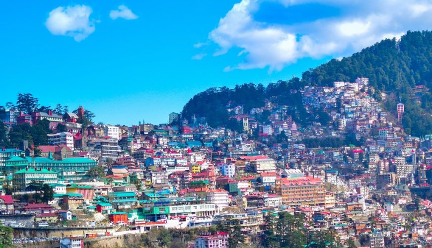 Panoramic view of Shimla's colorful buildings sprawled across lush green hills under a bright blue sky.